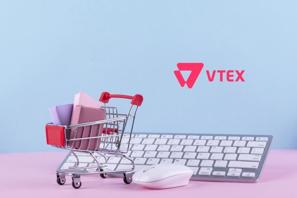 Carrinho de compras em miniatura sobre um teclado de computador, com pacotes coloridos e o logotipo da VTEX ao fundo, representando o e-commerce e as facilidades da plataforma VTEX IO.